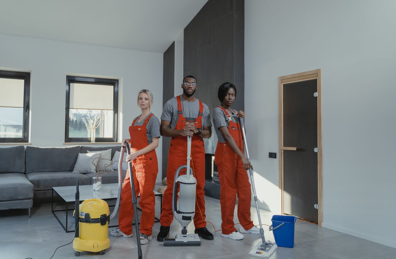 heros-img Diverse cleaning team in red uniforms ready to sanitize a modern living room with vacuum and mop.